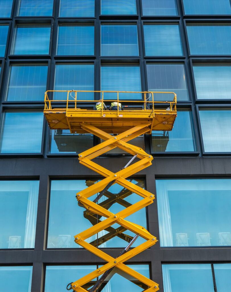 Yellow scissor lift elevated work platform making repairs to skyscraper windows.